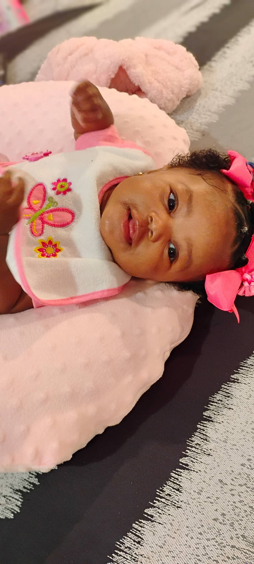 Jehlani is registered to the contest to win money with this photo: adorable, baby, bib, butterfly, child, closeup, curly_hair, cushion, cute, flower, headwear, indoors, infant, lying_down, pink, pink_bow, portrait, skin, smile, soft_texture