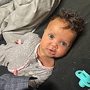 Nova is registered to the contest to win money with this photo: baby, infant, curly_hair, blue_eyes, pacifier, blanket, sleepwear, lying_down, indoor, cute, child, person, face, expression, soft_lighting, cozy, pink_trim, fabric, resting, portrait