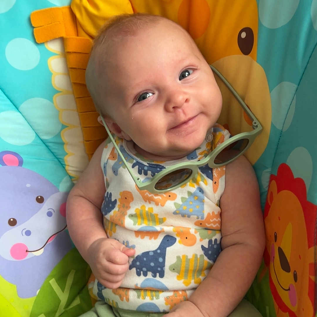 August joined the competition — help win amazing prizes! animal_print, baby, bright_colors, chair, child, colorful, cute, dinosaur_pattern, face, green_shorts, happy, indoors, infant, person, playful, portrait, seated, smiling, sunglasses, toy