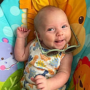 August is registered to the contest to win money with this photo: baby, infant, smiling, sunglasses, dinosaur_print, colorful, seat, animal_theme, playful, child, cute, happy, clothing, green_shorts, indoor, portrait, toy, hand, face, expression