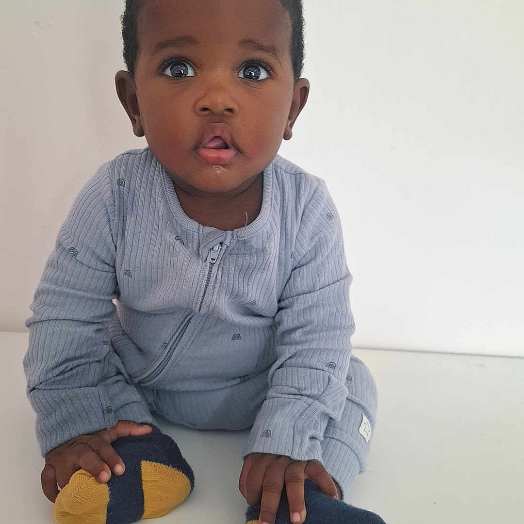 Munashe is registered to the contest to win money with this photo: baby, blue_outfit, child, closeup, clothing, curious, cute, expression, face, feet, hands, indoor, infant, person, portrait, sitting, socks, table, white_background, young_child