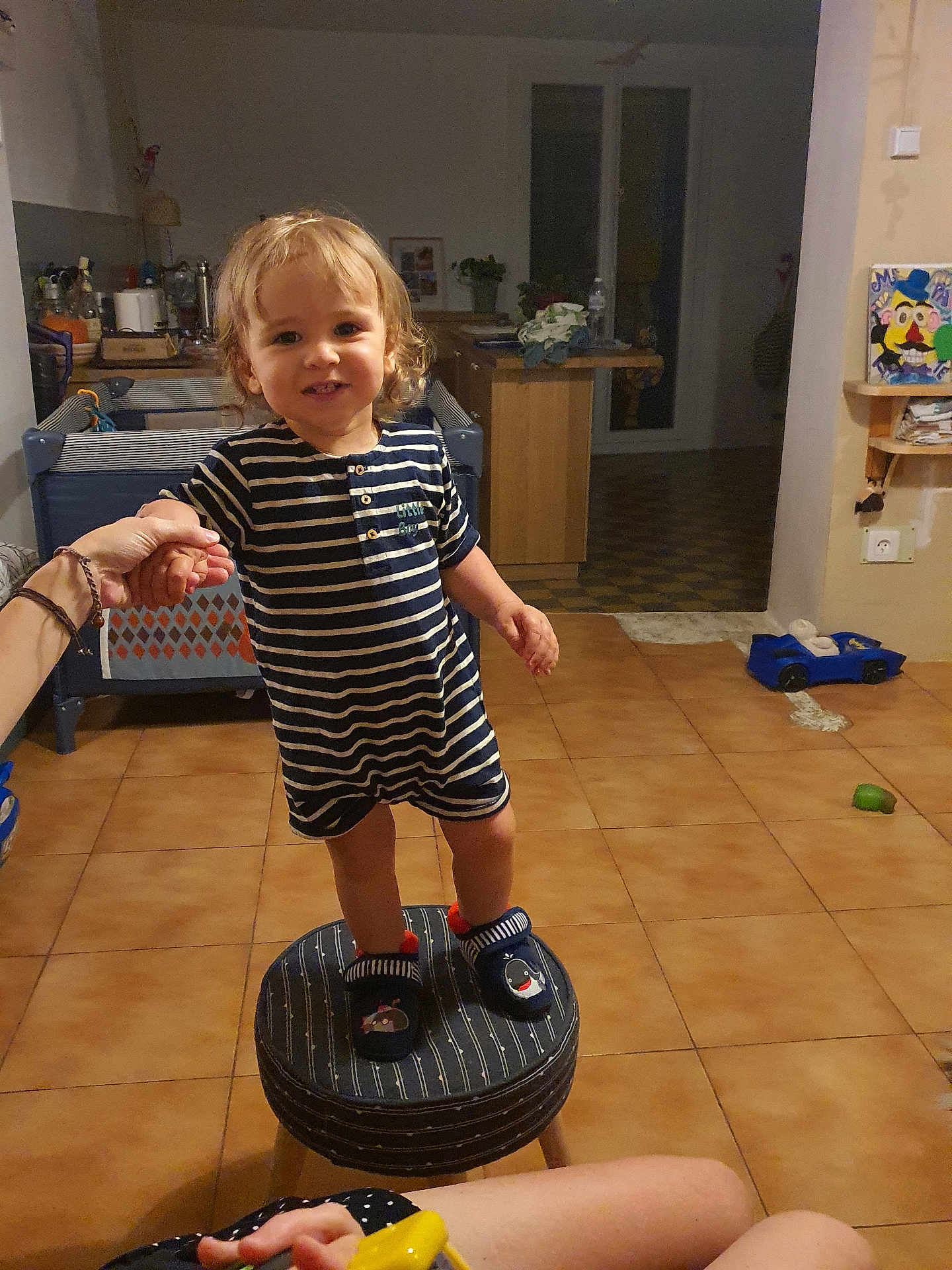 Arthur a rejoint le concours — aidez-le/la à gagner de superbes lots ! toddler, child, striped_pajamas, slippers, stool, hand_holding, indoor, kitchen, tile_floor, toy_car, furniture, person, smile, curly_hair, home, warm_lighting, adult_hand, background, cabinet, plant