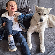 Tiago a rejoint le concours — aidez-le/la à gagner de superbes lots ! child, boy, dog, husky, pet, couch, blanket, denim_jacket, white_shirt, blue_pants, sneakers, socks, indoor, smiling, relaxed, companionship, furniture, home, animal, portrait