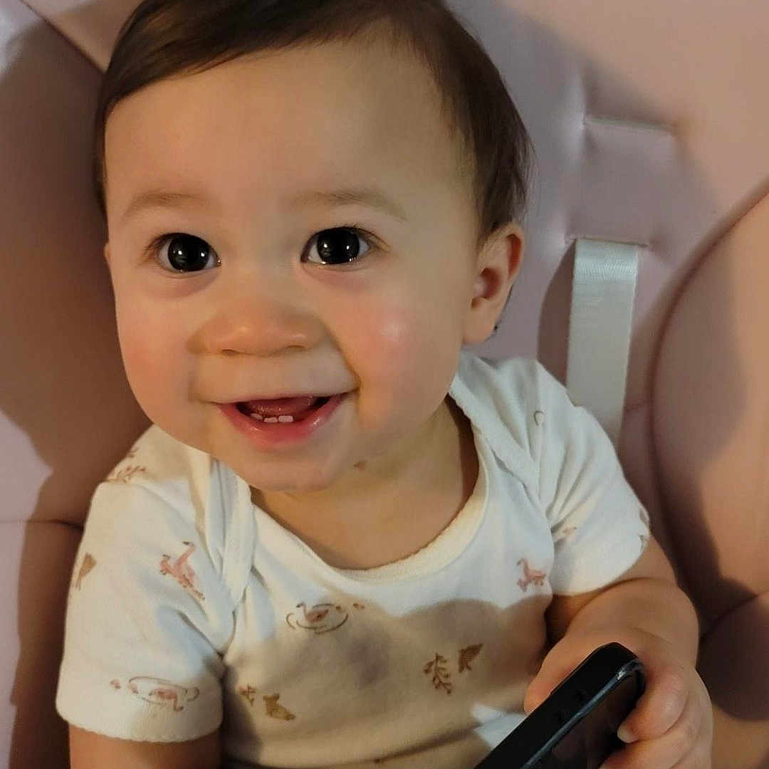 Ashlee is registered to the contest to win money with this photo: baby, child, cute, face, hand, happy, high_chair, indoor, infant, looking_up, person, phone, pigtails, playful, portrait, seated, smartphone, smiling, white_shirt, young
