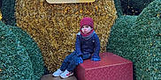 Micheal is registered to the contest to win money with this photo: child, winter_clothing, hat, gloves, scarf, gift_boxes, decorative_lights, greenery, outdoor, holiday_decoration, person, urban, building, sidewalk, cold_weather, festive, seasonal, expression, sitting, urban_outdoor