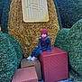 child, winter_clothing, hat, gloves, scarf, gift_boxes, decorative_lights, greenery, outdoor, holiday_decoration, person, urban, building, sidewalk, cold_weather, festive, seasonal, expression, sitting, urban_outdoor