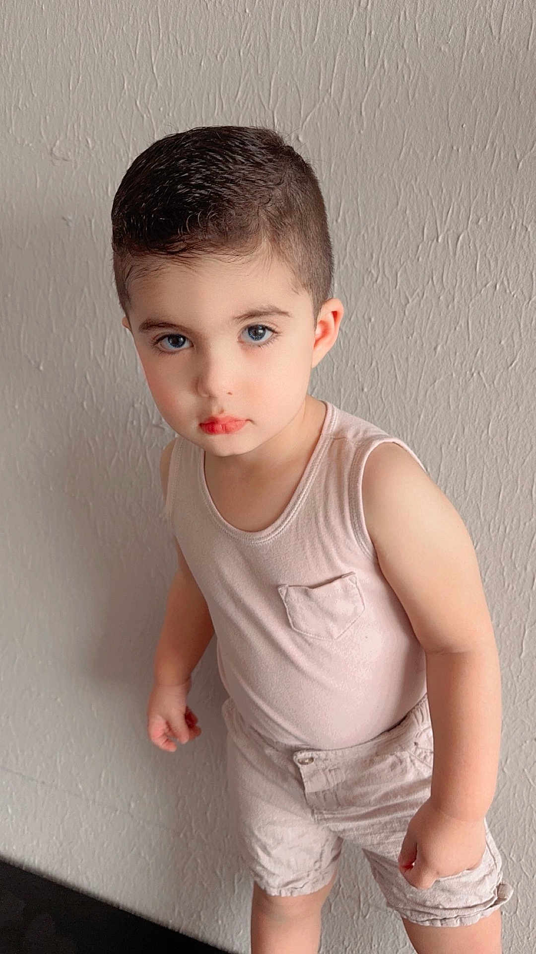 Micheal is registered to the contest to win money with this photo: child, toddler, boy, blue_eyes, short_hair, standing, light_clothing, sleeveless_shirt, shorts, indoor, wall, texture, casual, portrait, cute, serious_expression, person, skin, young, fashion