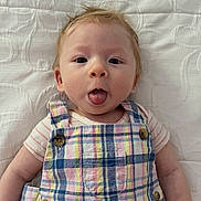Maximillian is registered to the contest to win money with this photo: baby, child, tongue_out, plaid, colorful_clothing, infant, cute, text_overlay, question, white_background, face, portrait, young_child, expression, lying_down, buttoned_overalls, short_sleeves, human, skin, funny
