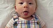 Maximillian is registered to the contest to win money with this photo: baby, child, tongue_out, plaid, colorful_clothing, infant, cute, text_overlay, question, white_background, face, portrait, young_child, expression, lying_down, buttoned_overalls, short_sleeves, human, skin, funny