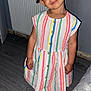 child, barefoot, striped_dress, curly_hair, wooden_floor, radiator, toy, indoor, playful_expression, person, smiling, standing, home, cute, colorful_clothing, buttoned_dress, young_girl, barefoot_child, portrait