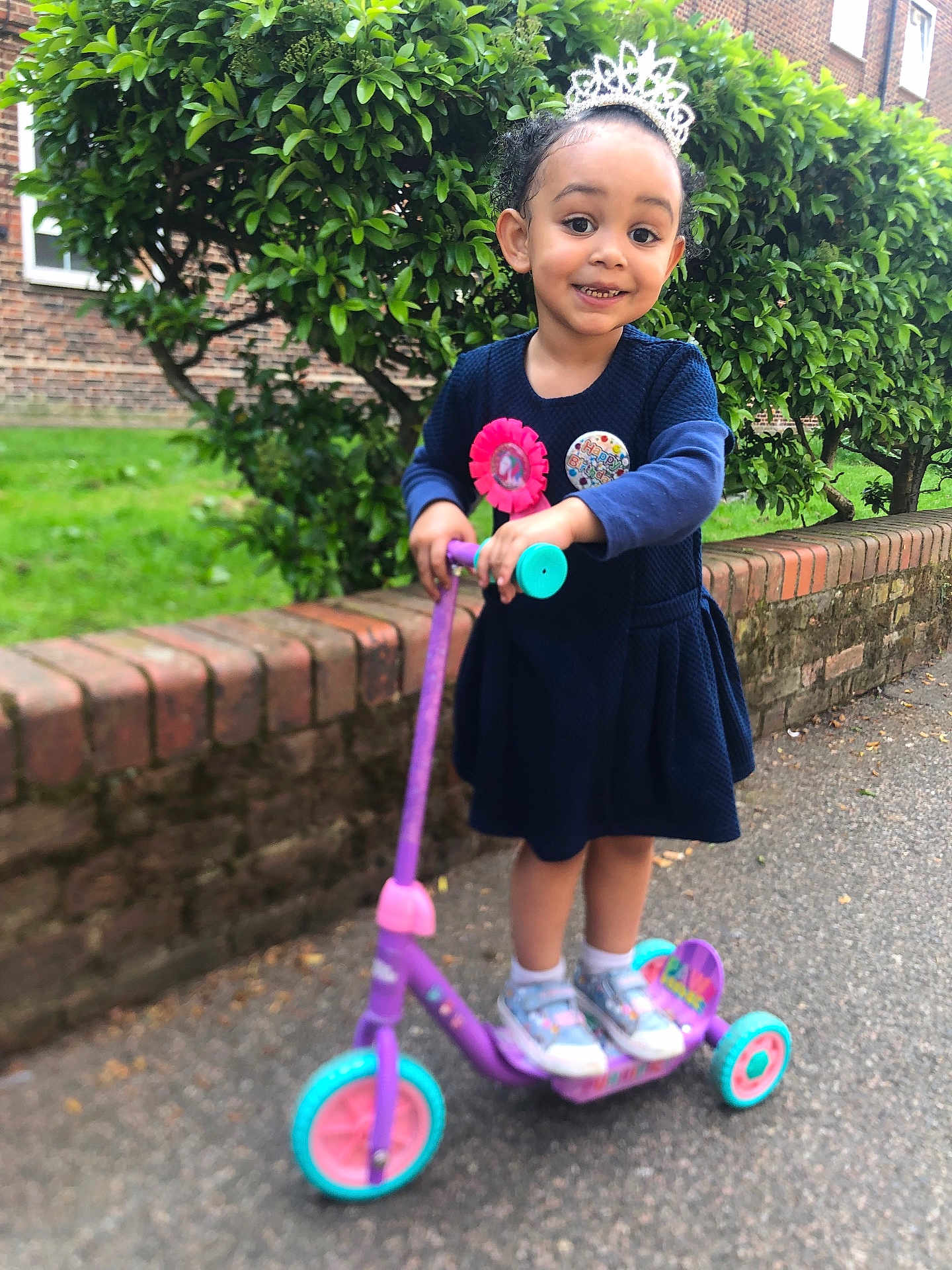 Krystal is registered to the contest to win money with this photo: child, girl, scooter, tiara, birthday_badge, outdoor, sidewalk, brick_wall, greenery, smiling, blue_dress, shoes, happy, playful, person, young_child, nature, celebration, fun, daytime