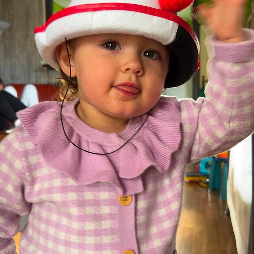 Iris participe au concours pour gagner de l'argent avec cette photo : buttoned_cardigan, child, cute, face, furniture, hat, home_interior, indoor, person, playful, pom_poms, portrait, purple_cardigan, remote_control, smiling, toddler, tongue_out, waving, wooden_floor, young_child