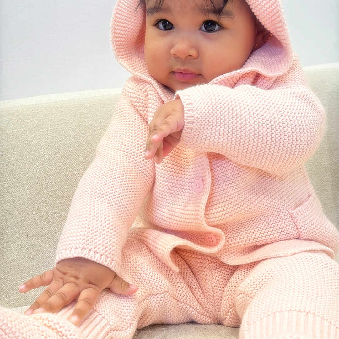 Aliana is registered to the contest to win money with this photo: baby, child, pink_clothing, hood, knitted, sitting, couch, cute, adorable, face, hand, footwear, indoors, portrait, young_child, cozy, soft_texture, warm_clothing, infant, expression