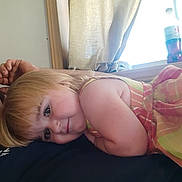 Emily is registered to the contest to win money with this photo: child, toddler, blonde_hair, smiling, indoor, window, curtain, bottle, person, fabric, clothing, face, arm, head, skin, light, sitting, resting, casual, portrait