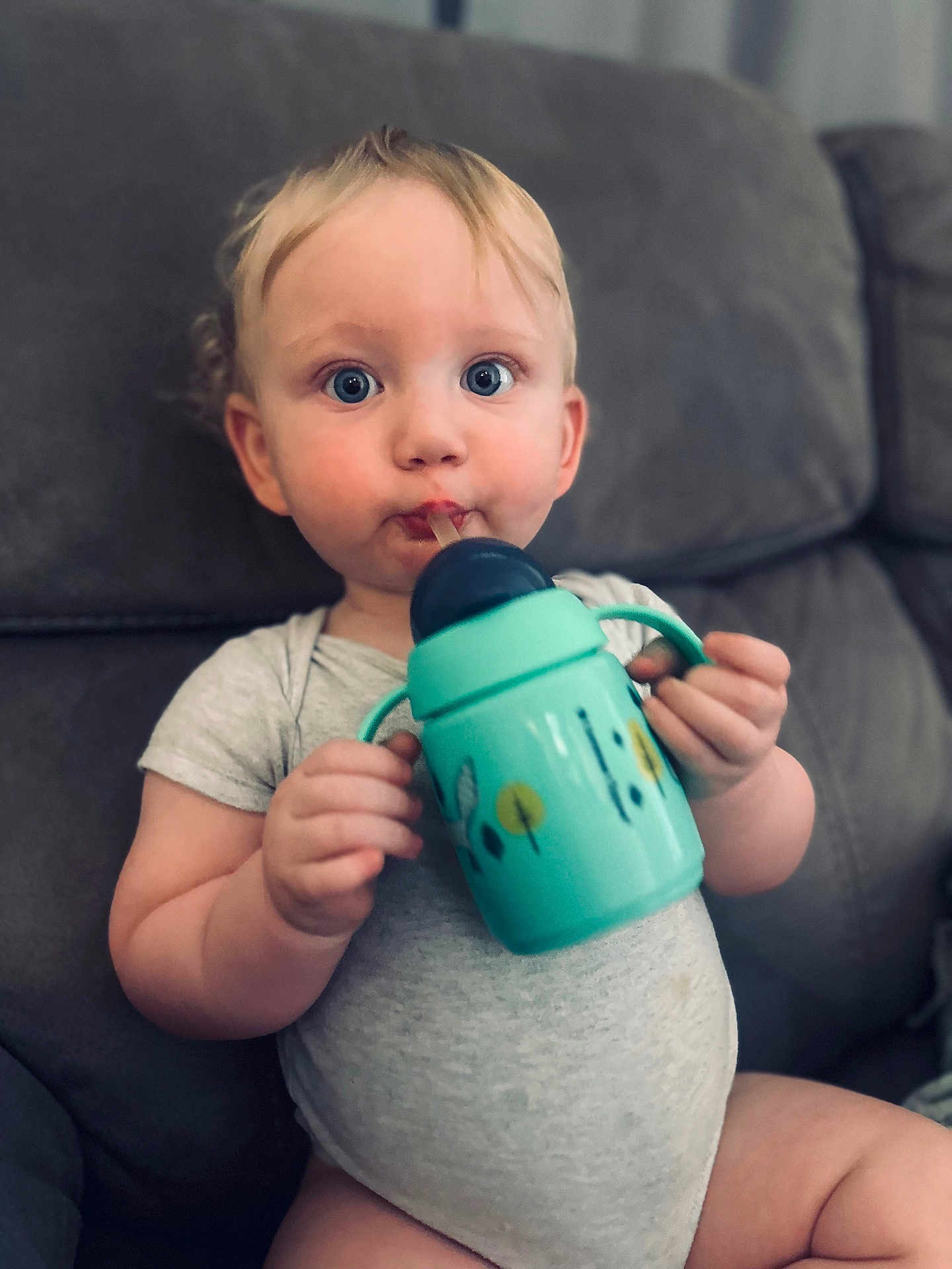 Oscar is registered to the contest to win money with this photo: baby, child, sippy_cup, blue_eyes, blond_hair, couch, gray_couch, onesie, sitting, indoor, drinking, face, hand, expression, cute, person, infant, young_child, furniture, home