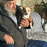 Otis is registered to the contest to win money with this photo: man, dog, beard, cap, jacket, sweater, couch, checkered, fireplace, brick, indoor, plant, photo_frame, hand, jeans, seated, pet, home, casual, portrait