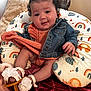 baby, child, denim_jacket, orange_outfit, curly_hair, hair_ties, white_sandals, flower_decorations, cushion, rainbow_pattern, animal_pattern, blanket, smiling, indoors, soft_lighting, cute, happy, person, seated, cozy
