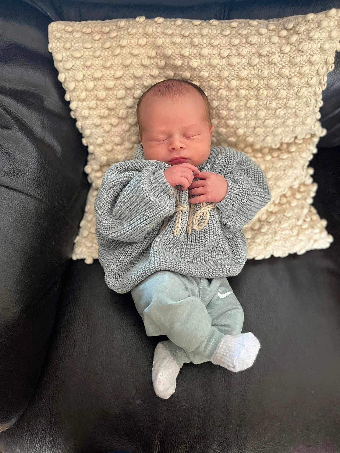 Albie joined the competition — help win amazing prizes! baby, newborn, sleeping, knitted_sweater, pants, white_socks, pillow, textured_pillow, leather_couch, indoor, cozy, cute, child, infant, resting, small_hands, peaceful, soft_lighting, portrait, casual_clothing