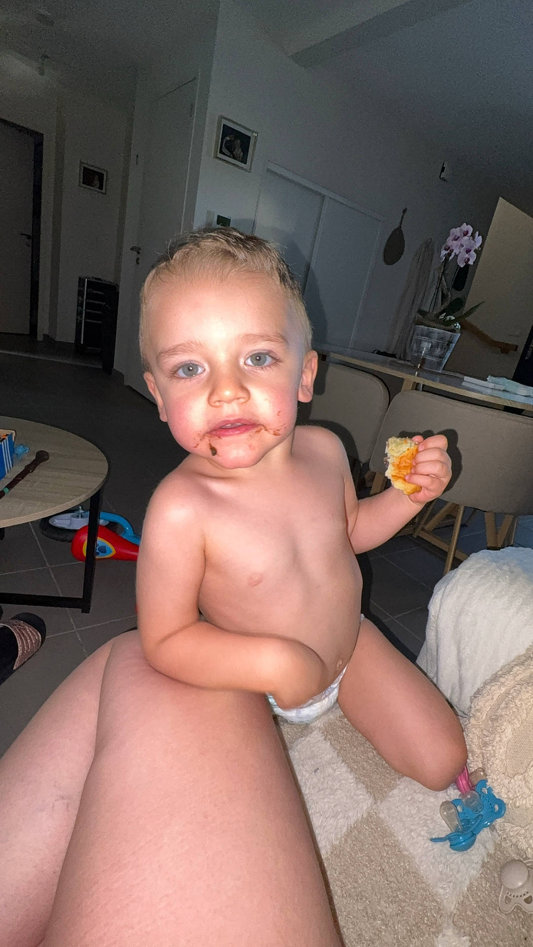 adult_leg, carpet, chair, child, cute, diaper, floor, happy, home, indoor, living_room, messy_face, pacifier, person, plant, skin, snack, table, toddler, toy