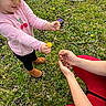 child, pink_shirt, cherries, curly_hair, flowers, grass, outdoor, nature, person, red_pants, hands, offering, wildflowers, greenery, casual_clothing, boots, spring, interaction, cute, playful
