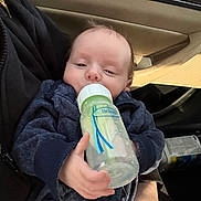 Isaiah joined the competition — help win amazing prizes! baby, infant, feeding_bottle, drinking, car_interior, car_door, seat, hand, parent_hand, jacket, clothing, face, eyes, mouth, feeding, close_up, portrait, cute, soft_lighting, relaxed