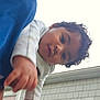baby, child, curly_hair, face, hand, outdoor, house, window, building, sky, person, portrait, clothing, blue_shirt, red_garment, young_child, looking_at_camera, close_up, daylight, casual
