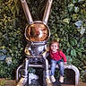 artistic, bench, bunny, child, concrete, cute, floor, greenery, helmet, indoor, jeans, leaves, plant_wall, pose, red_sweater, reflective, silver, smile, sneakers, statue