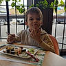 child, boy, sushi, food, restaurant, table, window, plants, indoor, meal, chopsticks, plate, car, glass, curious, casual_clothing, sitting, daylight, young, person