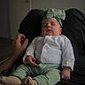 adult_hand, baby, blank_wall, bow, cozy, door, floor, green_pants, hand, headband, indoor, infant, lap, legs, newborn, portrait, sleeping, socks, soft_lighting, white_cardigan