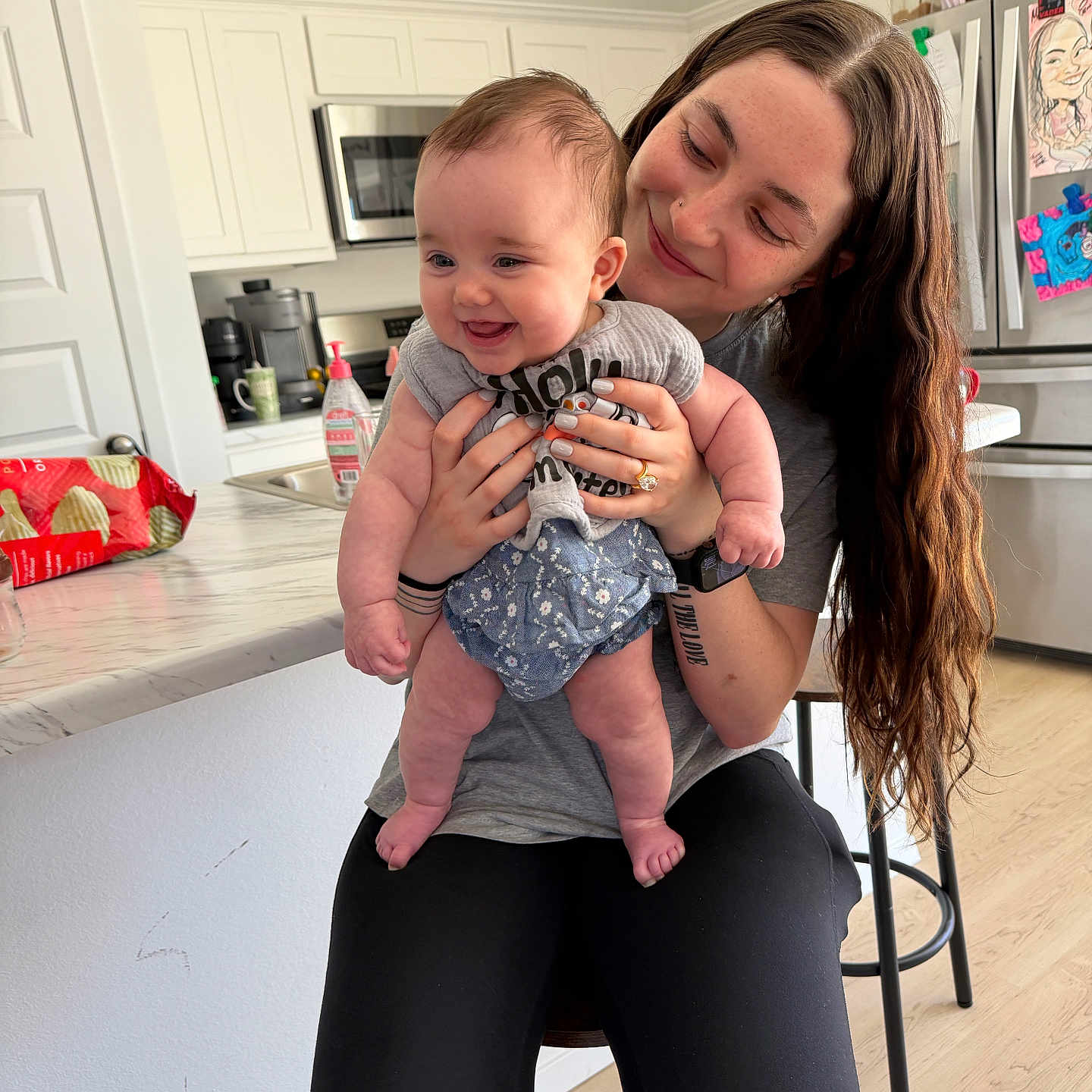 Sierra is registered to the contest to win money with this photo: baby, woman, kitchen, smile, holding, happy, person, long_hair, infant, casual_clothing, countertop, stool, refrigerator, indoor, family, portrait, home, cabinet, floor, appliance