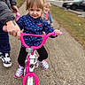 child, girl, bicycle, pink_bike, pigtails, adult_hand, outdoor, sidewalk, casual_clothing, footwear, support, learning, play, parked_car, background, daylight, determined, person, two_children, road