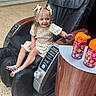 child, girl, massage_chair, black_chair, bows, sandals, water_bottles, table, indoor, floor_tiles, happy, smiling, short_sleeve_dress, light_dress, hair_accessories, person, furniture, relaxation, public_space, cute