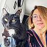 cat, tuxedo_cat, black_cat, white_chest, pet, animal, woman, person, selfie, portrait, indoors, home, glasses, brown_hair, striped_shirt, golden_eyes, whiskers, paws, sitting, close_up