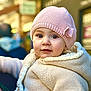 baby, child, knit_hat, pink_hat, bow, cream_jacket, knit_jacket, face, portrait, outdoor, curious, warm_clothing, person, cute, young_child, urban_background, soft_focus, closeup, winter_wear, innocent