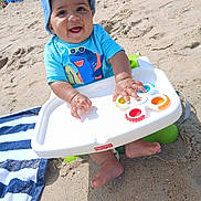 Major is registered to the contest to win money with this photo: baby, child, beach, sand, sunhat, blue_shirt, toy, fidget_spinner, fisher_price, smiling, happy, seated, sunny, outdoor, footwearless, towel, striped_towel, playful, summer, infant