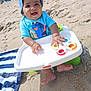 baby, child, beach, sand, sunhat, blue_shirt, toy, fidget_spinner, fisher_price, smiling, happy, seated, sunny, outdoor, footwearless, towel, striped_towel, playful, summer, infant
