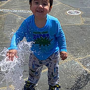 Emilio joined the competition — help win amazing prizes! child, toddler, water, splash, fountain, blue_shirt, dinosaur_print, pajama_pants, shoes, wet_hair, smiling, outdoor, play, fun, sunny, ground, pavement, happy, person, daytime