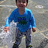 child, toddler, water, splash, fountain, blue_shirt, dinosaur_print, pajama_pants, shoes, wet_hair, smiling, outdoor, play, fun, sunny, ground, pavement, happy, person, daytime