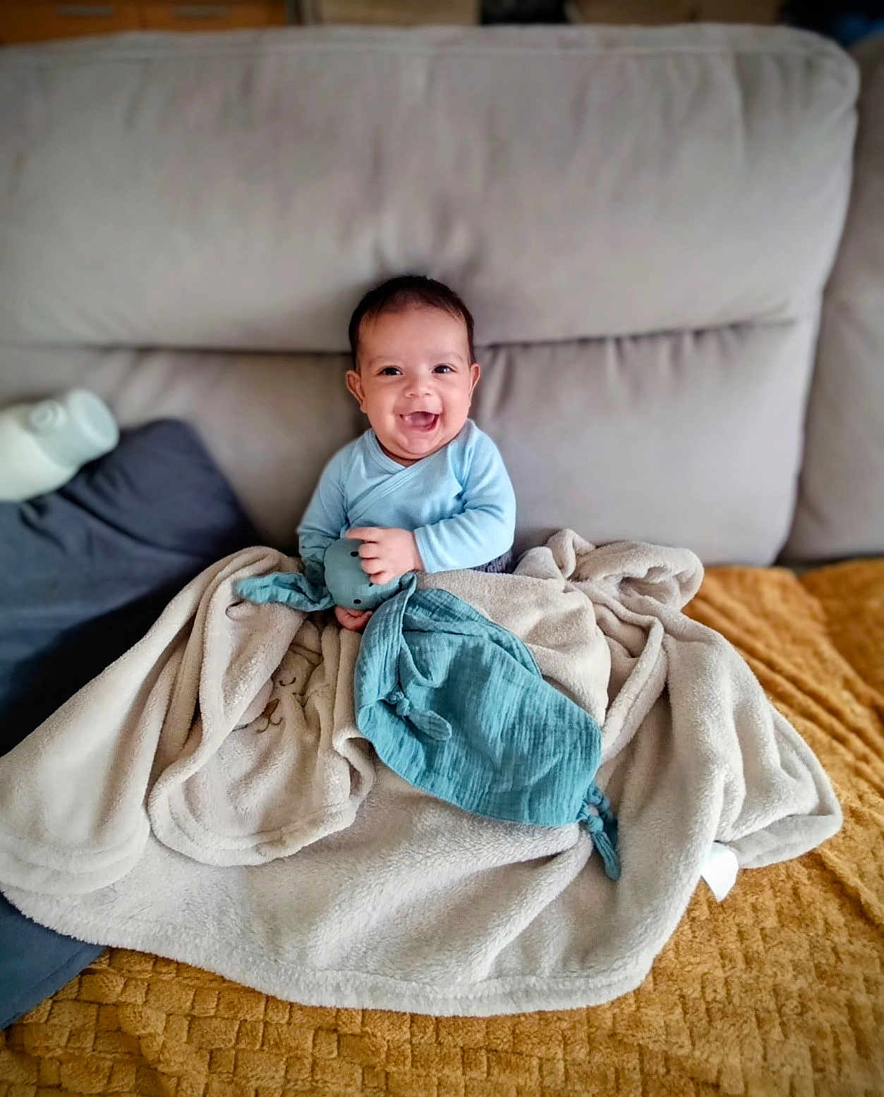 Naël participe au concours pour gagner de l'argent avec cette photo : baby, smiling, blue_clothing, blanket, toy, couch, indoor, child, happy, soft_toy, cozy, seated, face, infant, home, comfort, plush, fabric, person, cute