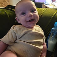 Atlus is registered to the contest to win money with this photo: baby, smile, infant, couch, green_couch, bottle, milk_bottle, crate, purple_crate, beige_clothing, ribbed_clothing, happy, indoors, person, child, face, seated, baby_bottle, cozy, home