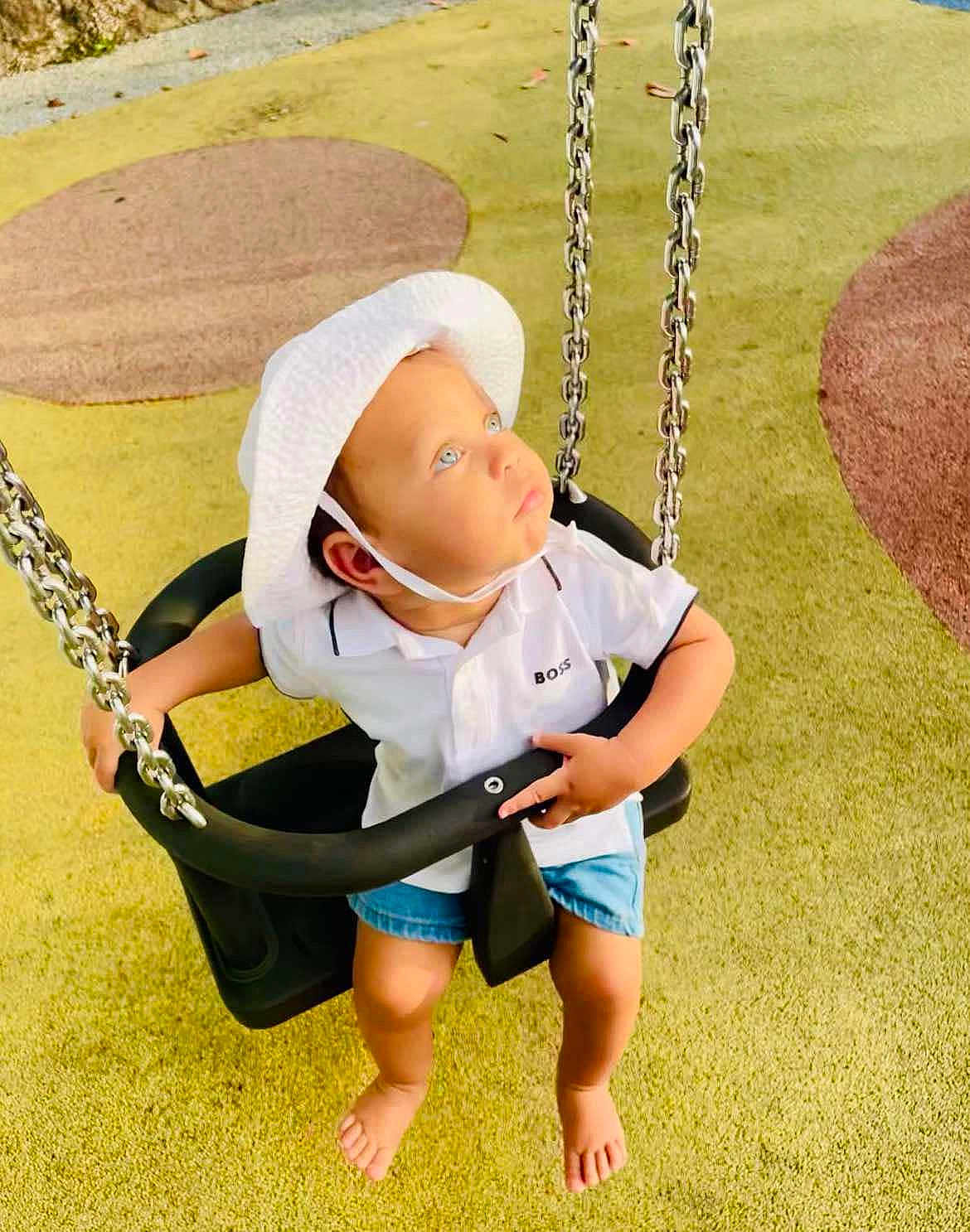 Ilyass a rejoint le concours — aidez-le/la à gagner de superbes lots ! toddler, swing, playground, child, hat, white_shirt, shorts, barefoot, chains, yellow_surface, looking_up, outdoor, sunlight, curious, baby_swing, play_area, childrens_clothing, summer, safety_seat, bright_eyes