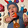 baby, blanket, blue_clothing, bunny_ears, child, colorful, curious_expression, cute, face, hand, hat, indoor, infant, lying_down, orange_toy, plastic_eggs, playtime, red_ears, soft_texture, toy