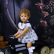 ève Horn participe au concours pour gagner de l'argent avec cette photo : child, toddler, curly_hair, dress, toy_car, black_toy_car, white_socks, white_shoes, bow, bracelet, teddy_bear, heart_shape, wooden_frame, roses, floral_backdrop, fur_rug, indoor, portrait, smiling, cute