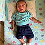 baby, child, smiling, lying_down, pillow, blanket, heart_pattern, blue_shirt, shorts, socks, indoors, cute, happy, person, infant, cozy, soft, playful, young, resting