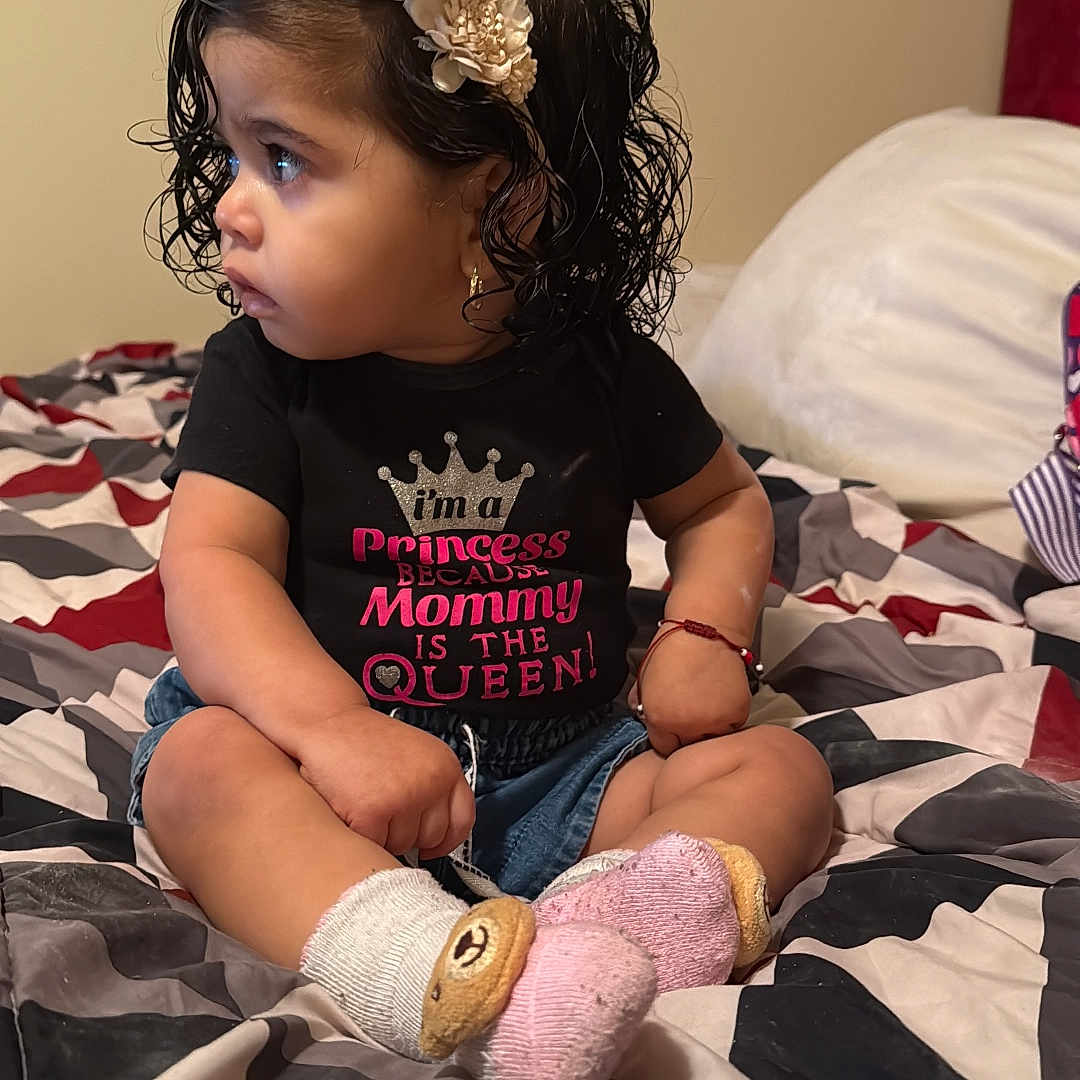 Estefani joined the competition — help win amazing prizes! bedroom, bedspread, black_shirt, bracelet, child, cross_legged, curly_hair, cute, earrings, expression, girl, headband, indoor, pink, portrait, shirt, sitting, socks, text_on_clothing, toddler