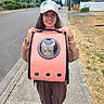 Astro joined the competition — help win amazing prizes! person, cat, pet_carrier, sidewalk, street, suburban, outdoor, daytime, smiling, cap, trees, grass, house, asphalt, casual_clothing, curious_cat, holding, walking, nature, happy