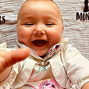 Arabella is registered to the contest to win money with this photo: baby, smiling, reaching_hand, floral_dress, cardigan, blanket, face, happy, infant, portrait, child, cute, closeup, skin, head, person, baby_girl, expression, lying_down, clothing