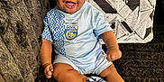 Kazi joined the competition — help win amazing prizes! baby, blue_clothing, child, couch, cushion, cute, fabric_texture, footwear, happy, indoor, infant, joyful, patterned_cushion, person, shorts, sitting, smiling, sock, text_on_clothing, young_child