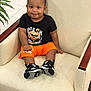 Kazi is registered to the contest to win money with this photo: armchair, baby, black_tshirt, child, cute, fashion, floor, furniture, home, indoor, leisure, orange_shorts, person, plant, portrait, sitting, smiling, sneakers, toddler, wood