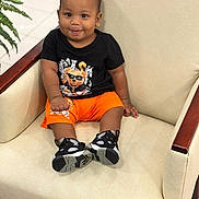 Kazi joined the competition — help win amazing prizes! armchair, baby, black_tshirt, child, cute, fashion, floor, furniture, home, indoor, leisure, orange_shorts, person, plant, portrait, sitting, smiling, sneakers, toddler, wood